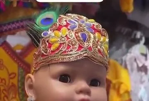 A baby doll modelling a yellow and red Krishna festive costume with a gold-embroidered, peacock-feather-topped crown.