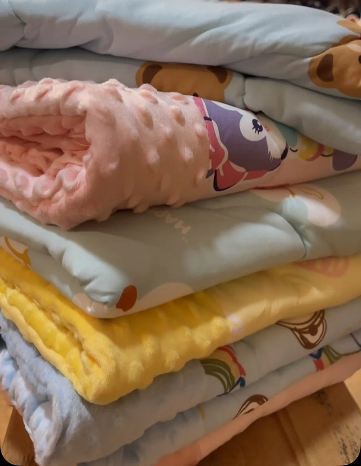 A stack of folded muslin baby blankets in pastel blue, pink and yellow, with cartoon bear and animal prints.